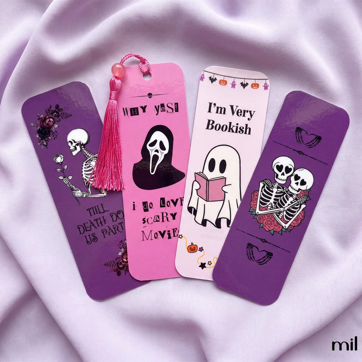 Four Halloween-themed paper bookmarks arranged on a light lavender satin fabric background. The bookmarks feature purple and pink colors with cartoon skeleton and ghost illustrations, plus text such as 'Till Death Do Us Party,' 'I Do Love Scary Movies,' 'I'm Very Bookish,' and 'Halloween.
