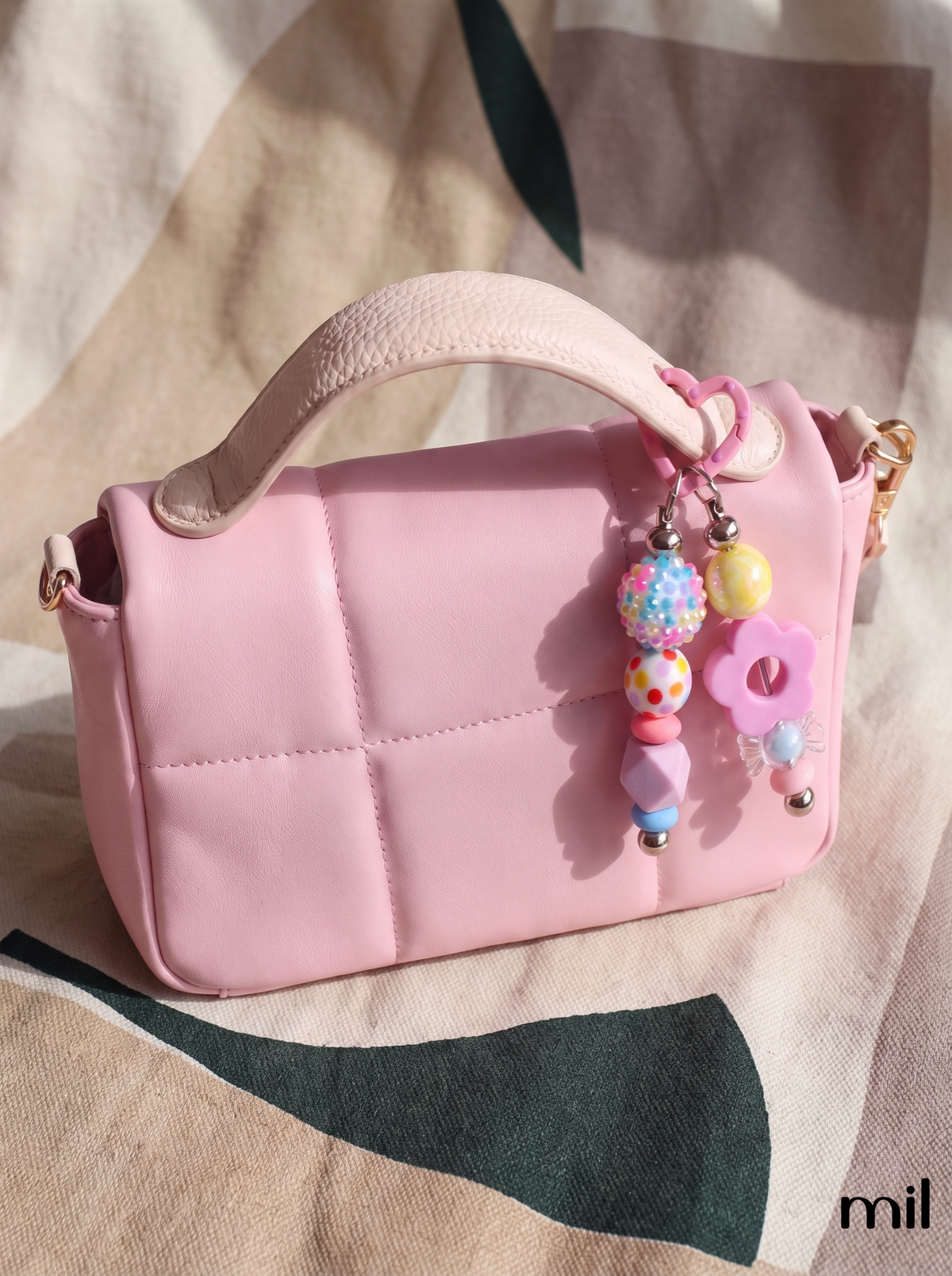 A pink quilted faux-leather handbag with a top handle, decorated with a colorful beaded keychain charm, resting on a beige and green patterned fabric background.