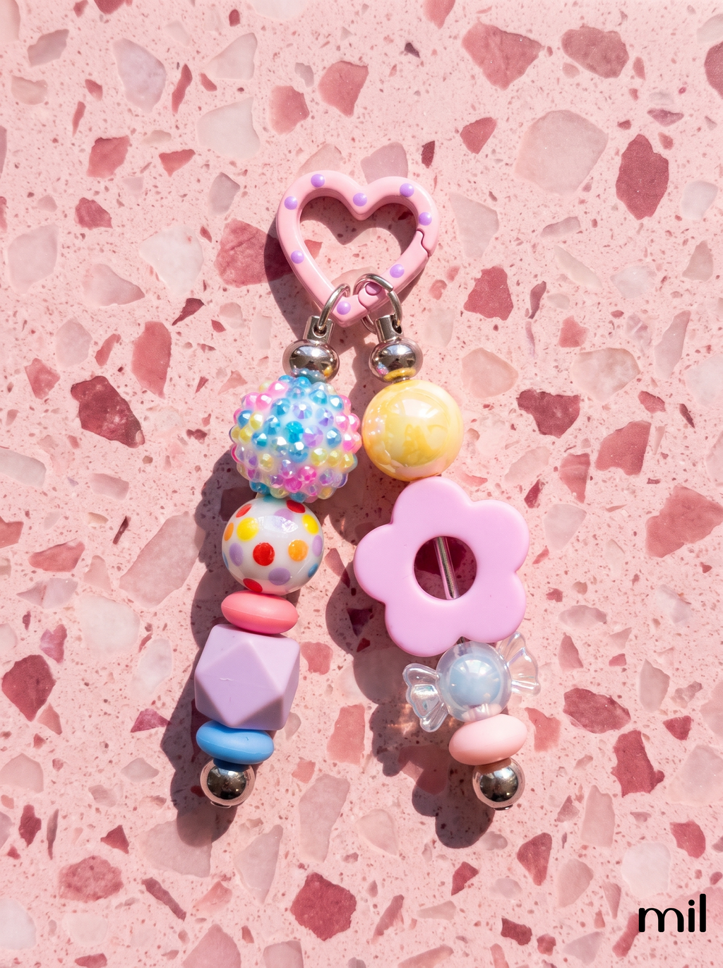 A pastel pink and multicolor beaded keychain charm with a heart-shaped ring, flower bead, and patterned beads, displayed on a pink terrazzo surface.