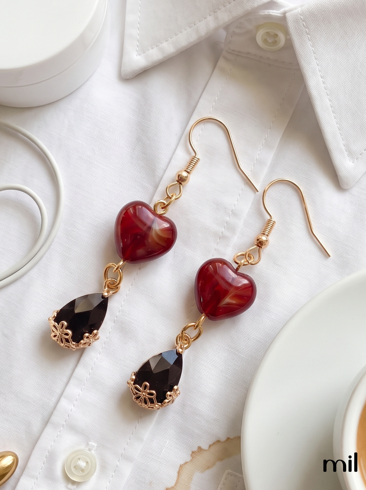 A pair of gold-tone dangle earrings with heart-shaped marbled red beads and teardrop-shaped dark brown beads, styled on a white collared shirt with coffee cups in the background.