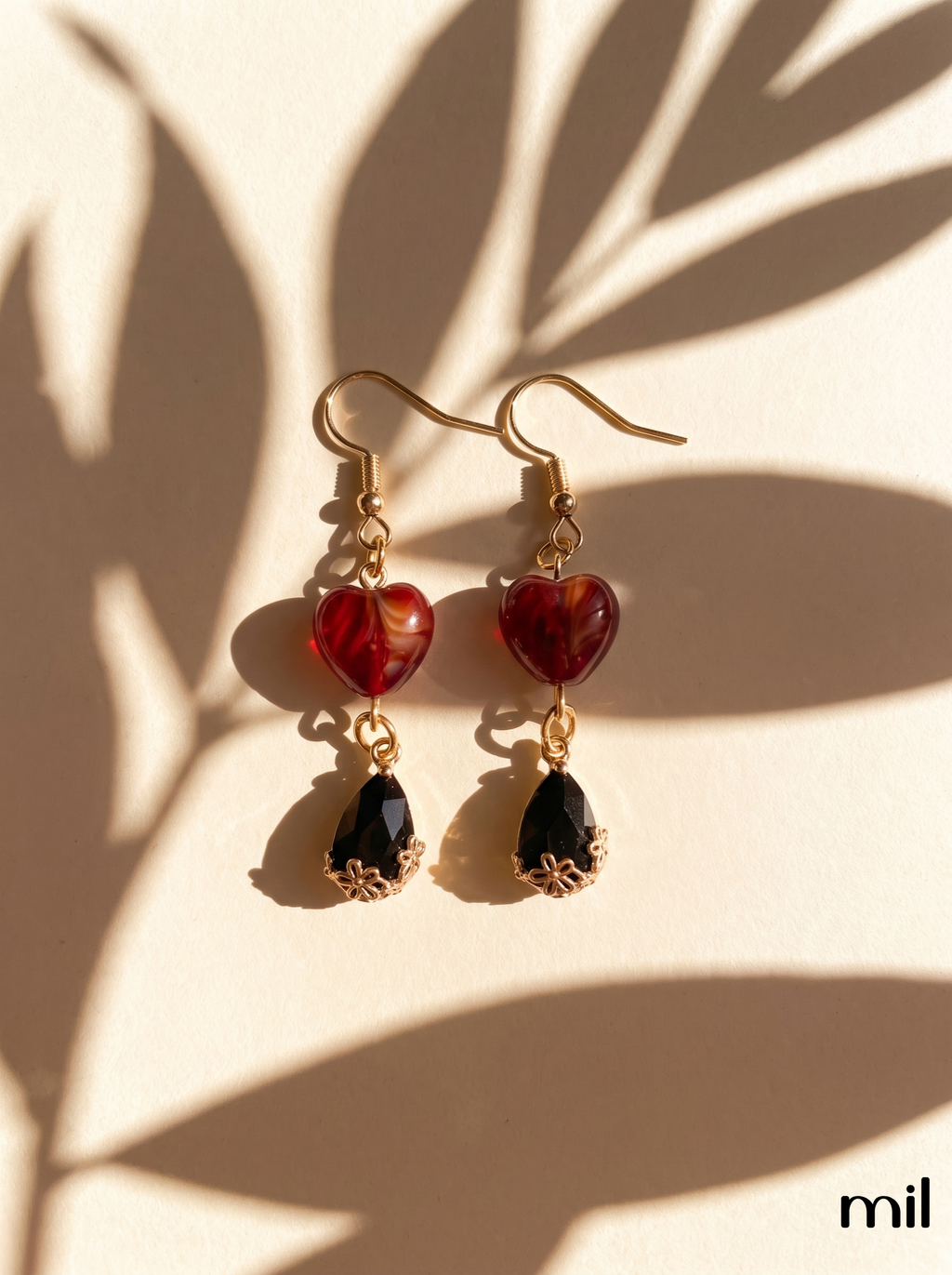 A pair of gold-tone drop earrings with heart-shaped red beads and teardrop-shaped black beads, displayed on a warm beige surface with leaf shadows.