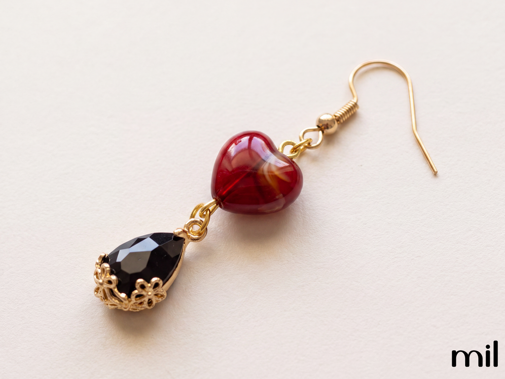 A pair of gold-tone dangle earrings featuring a glossy red heart-shaped bead and a faceted black teardrop gemstone in an ornate filigree setting, displayed on a light cream background.