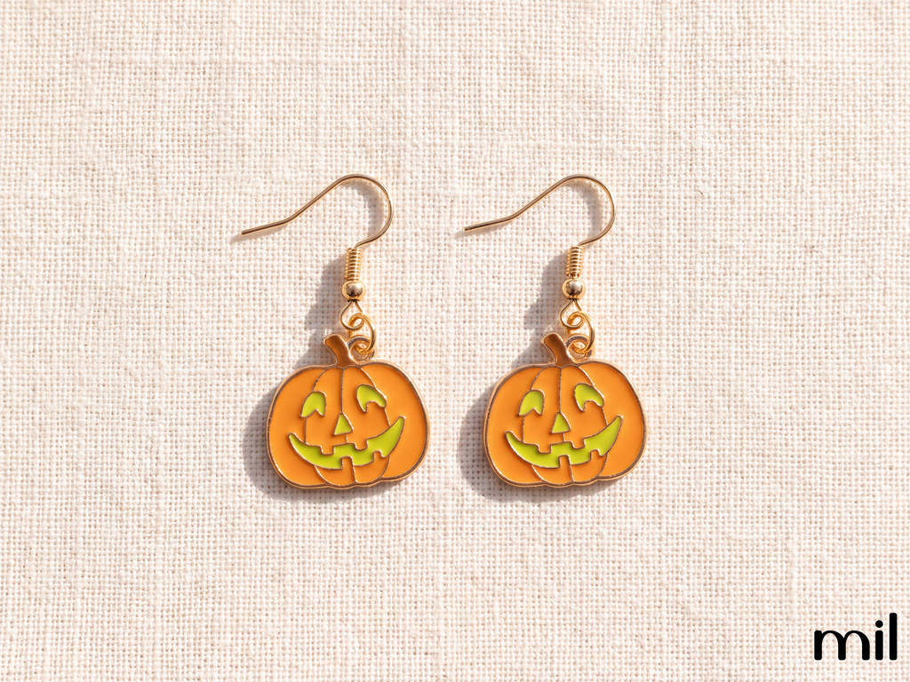 A pair of gold-tone dangle earrings featuring orange pumpkin charms with green jack-o'-lantern faces, displayed on a textured beige fabric background.