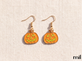 A pair of gold-tone dangle earrings featuring orange pumpkin charms with green jack-o'-lantern faces, displayed on a textured beige fabric background.