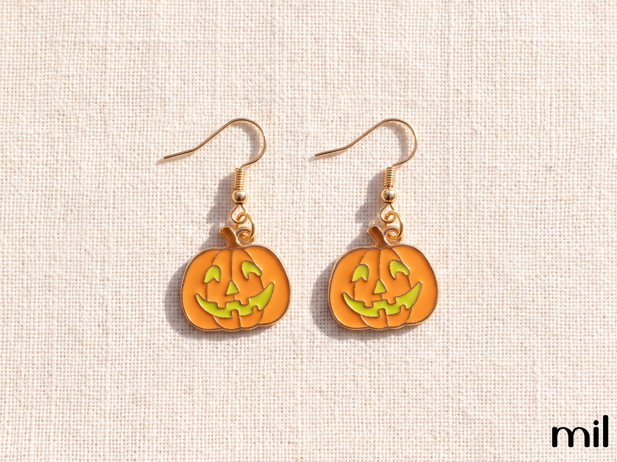A pair of gold-tone dangle earrings featuring orange pumpkin charms with green jack-o'-lantern faces, displayed on a textured beige fabric background.
