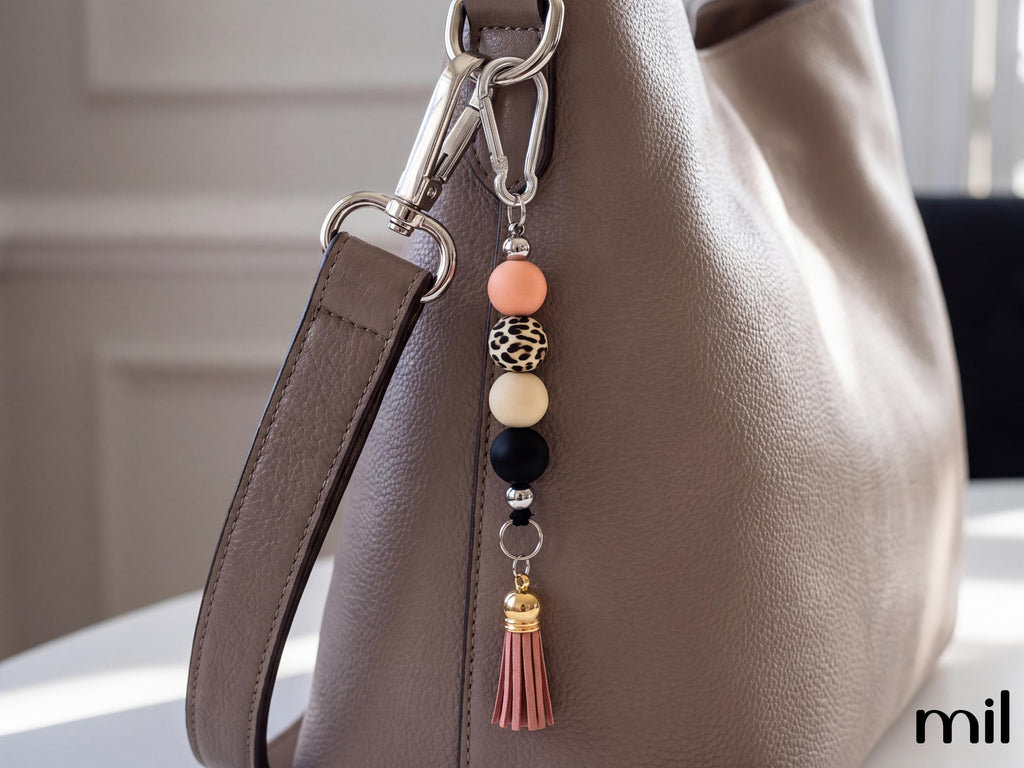 A close-up of a taupe leather shoulder bag with a decorative keychain charm featuring peach, leopard-print, cream, and black beads, a silver carabiner, and a pink tassel.