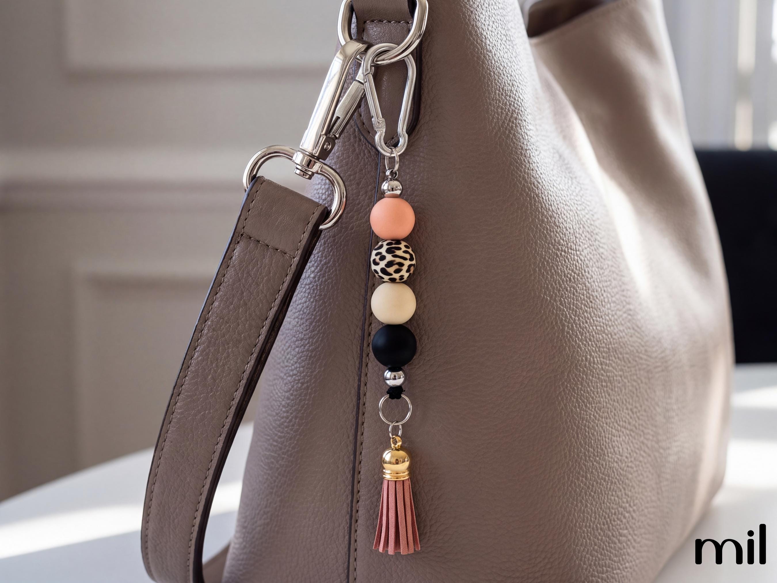A close-up of a taupe leather shoulder bag with a decorative keychain charm featuring peach, leopard-print, cream, and black beads, a silver carabiner, and a pink tassel.