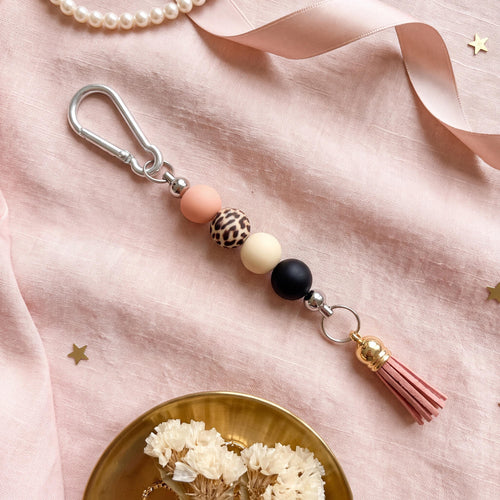 A beaded keychain charm with a silver carabiner clip, featuring pastel pink, leopard print, cream, and black beads, a pink tassel, and a gold-toned accent, styled on a pink satin fabric background.