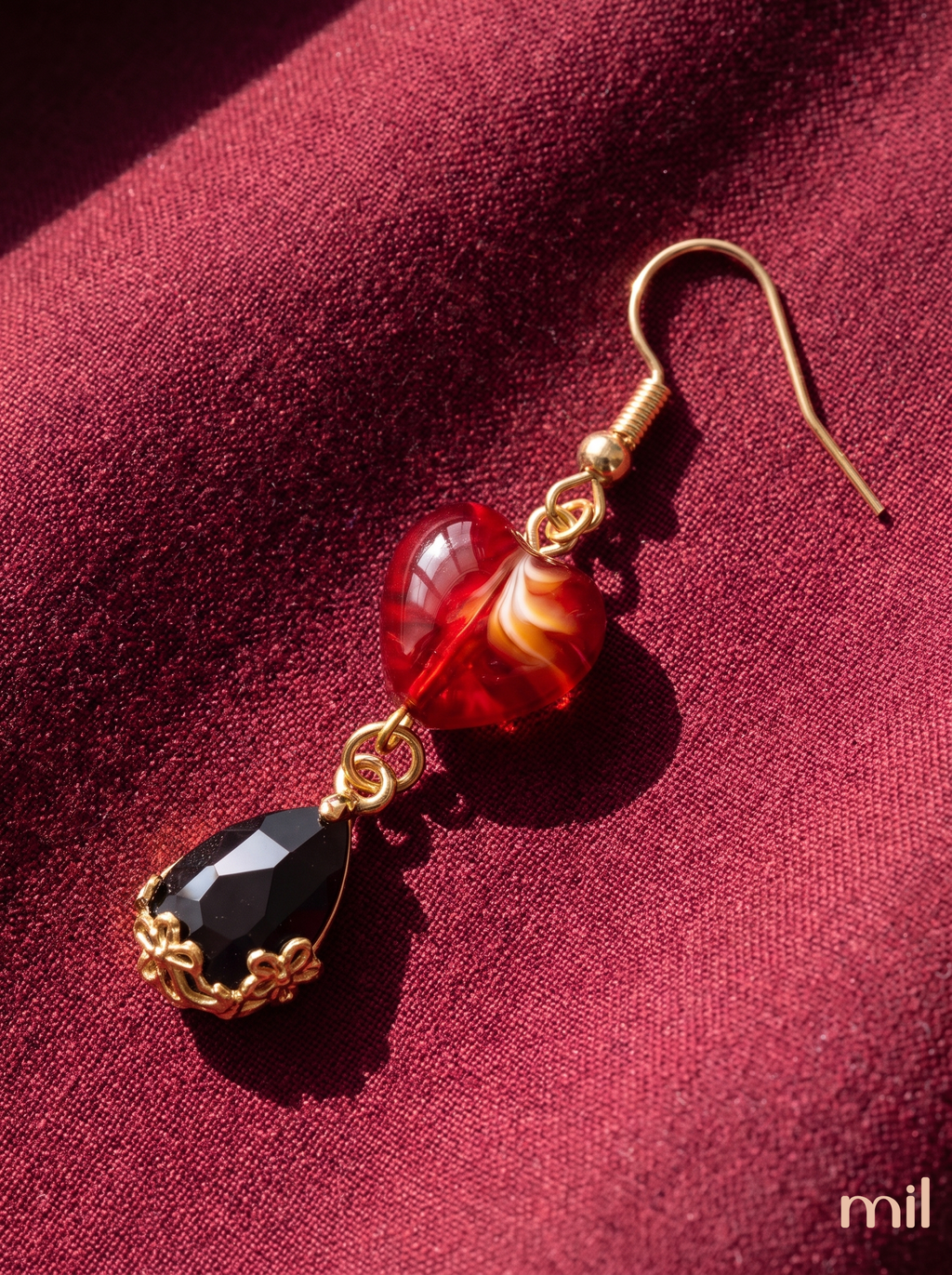 A single dangling earring with a gold-tone hook, a translucent red and orange marbled bead, and a faceted black teardrop stone set in ornate gold, photographed on a deep burgundy fabric background.