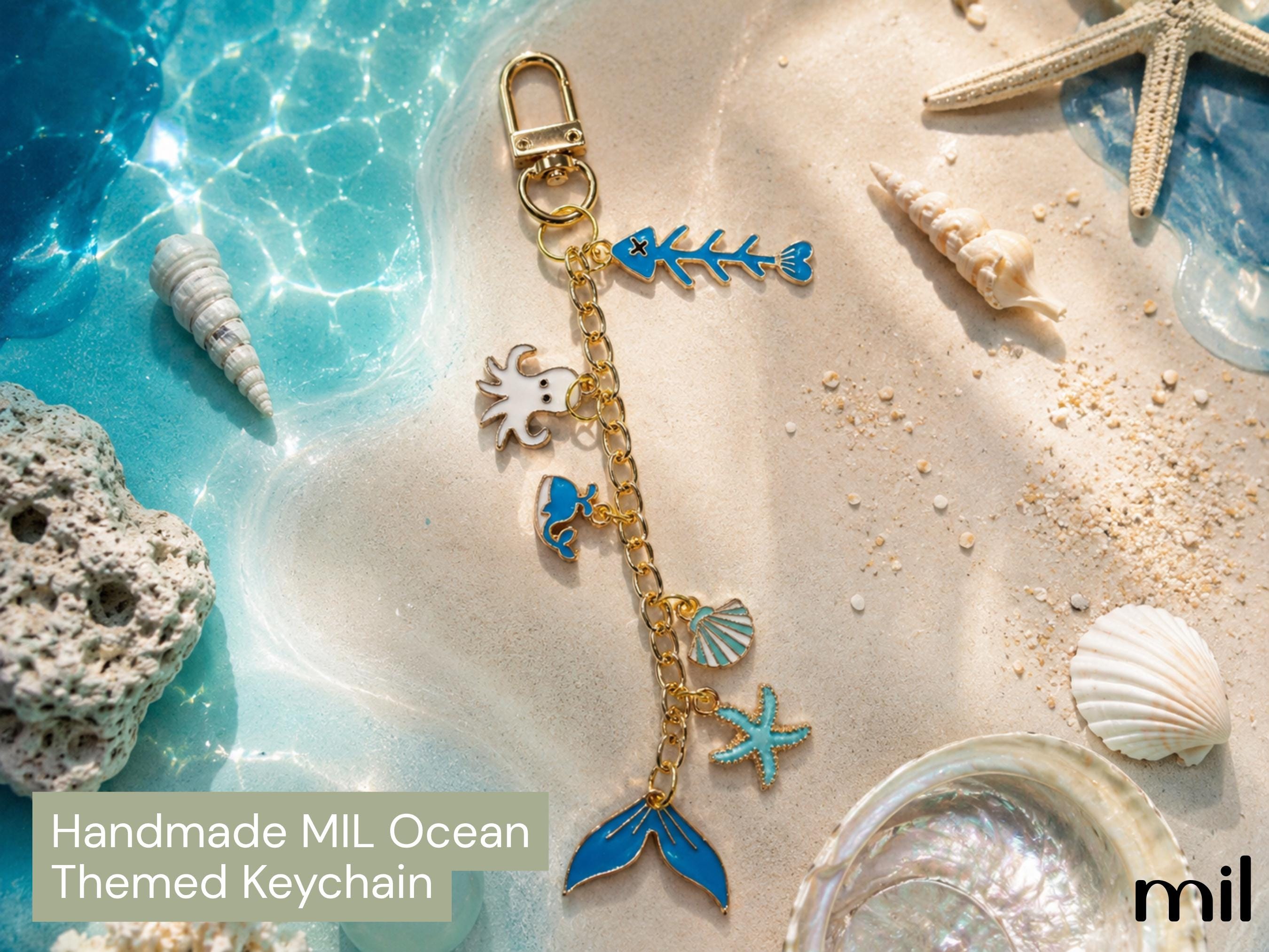A handmade ocean-themed keychain with gold-tone metal and blue enamel charms, displayed on a sandy beach background with seashells, starfish, and turquoise water. Text overlay reads 'Handmade MIL Ocean Themed Keychain' and the brand 'mil' appears in the bottom right corner.