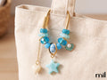 A close-up of a beige canvas tote bag with a decorative charm hanging from the top handle. The charm features gold-tone metal rings and chains, blue and light blue beads, a star-shaped pendant, a heart-shaped pendant, and a small shell charm.