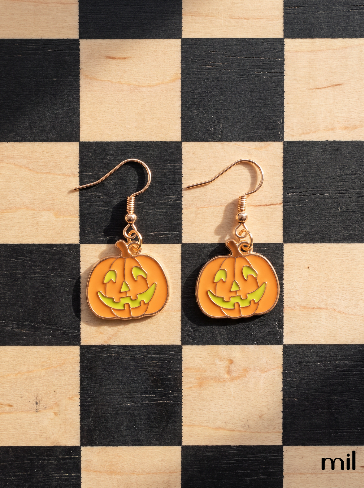 A pair of Halloween-themed drop earrings featuring orange jack-o'-lantern pendants with yellow facial details, suspended from rose gold hooks, displayed on a black and beige checkered wooden surface.