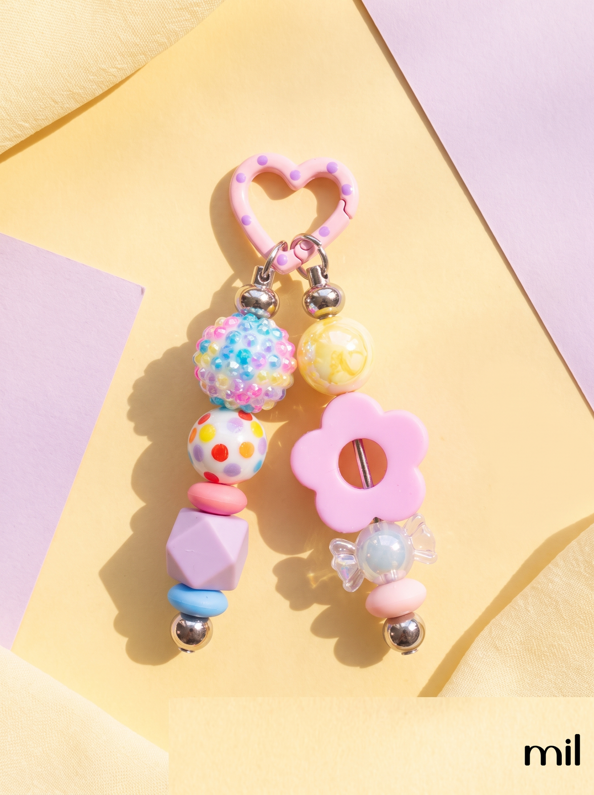A pastel pink and multicolor beaded keychain charm with a heart-shaped ring, flower-shaped bead, and colorful decorative beads, displayed on a pastel yellow and lavender background.