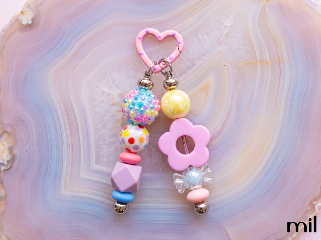 A pastel pink and multicolor beaded keychain or bag charm with a heart-shaped pink clasp, decorative beads, a flower-shaped pink bead, and small metallic bells, displayed on a pink and purple agate slice background.