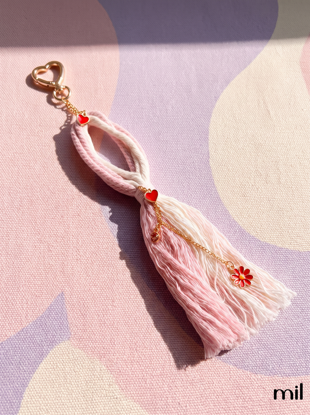 A pink and cream tassel keychain with gold-tone hardware, featuring small red heart and flower charms, displayed on a pastel patterned fabric background.
