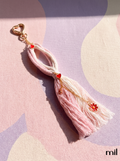 A pink and cream tassel keychain with gold-tone hardware, featuring small red heart and flower charms, displayed on a pastel patterned fabric background.