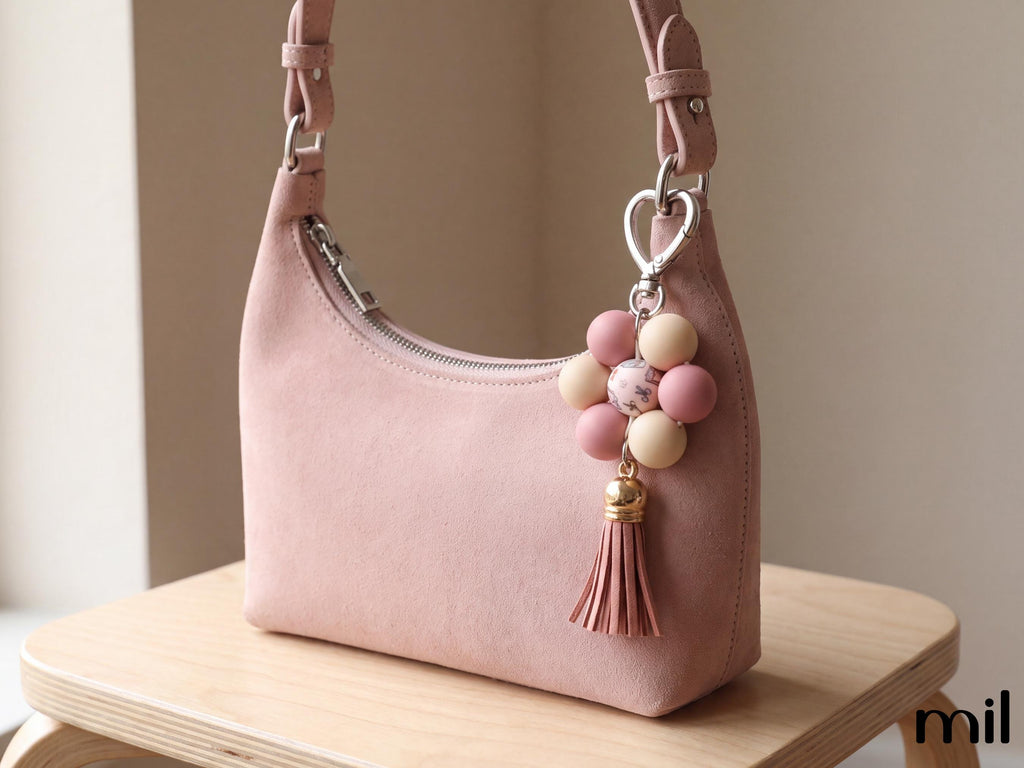 A dusty pink faux-leather shoulder bag with a floral bead keychain charm, resting on a light wood stool against a neutral beige background.