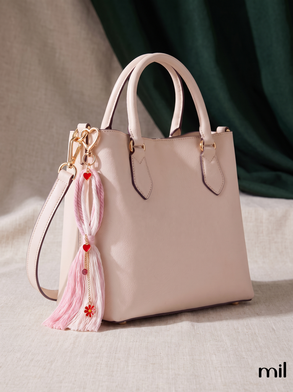 A light blush pink leather tote bag with double top handles, gold-tone hardware, and a decorative pink tassel charm with red hearts and a flower, shown on a draped beige fabric background with a dark green curtain.