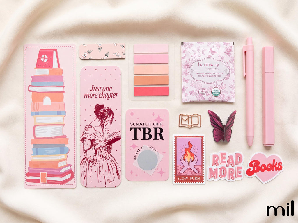 A coordinated set of pink stationery and book-themed accessories arranged on a soft cream fabric background. The set includes a tall bookmark with a stack of illustrated books, a narrow card with pink floral pattern, a stack of pink color swatches, a small packaged tea bag labeled 'harmony', two pink pens, a butterfly-shaped accessory, a stamp-style card, and a heart-shaped 'Read More Books' sticker.