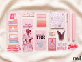 A coordinated set of pink stationery and book-themed accessories arranged on a soft cream fabric background. The set includes a tall bookmark with a stack of illustrated books, a narrow card with pink floral pattern, a stack of pink color swatches, a small packaged tea bag labeled 'harmony', two pink pens, a butterfly-shaped accessory, a stamp-style card, and a heart-shaped 'Read More Books' sticker.
