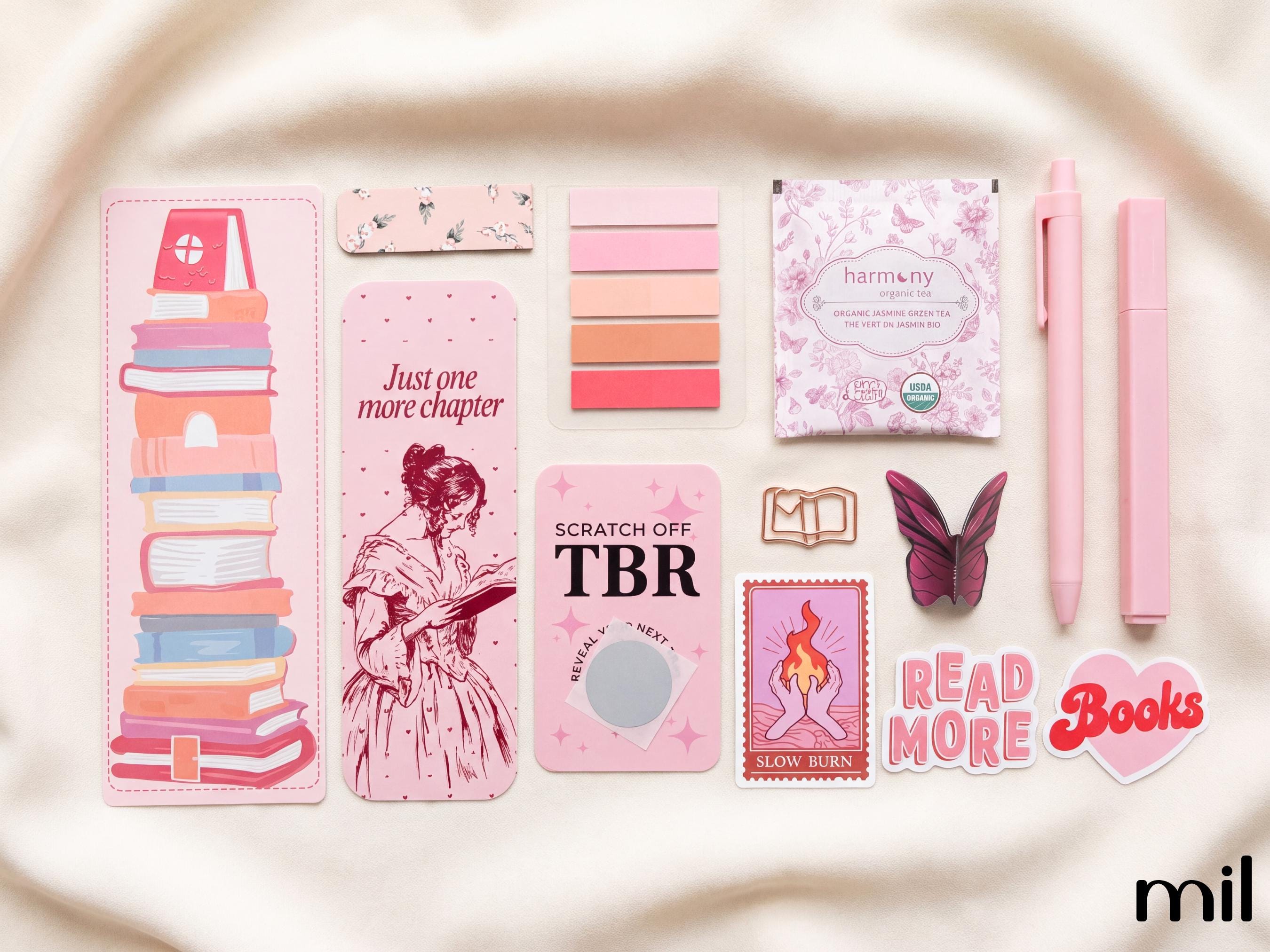 A coordinated set of pink stationery and book-themed accessories arranged on a soft cream fabric background. The set includes a tall bookmark with a stack of illustrated books, a narrow card with pink floral pattern, a stack of pink color swatches, a small packaged tea bag labeled 'harmony', two pink pens, a butterfly-shaped accessory, a stamp-style card, and a heart-shaped 'Read More Books' sticker.