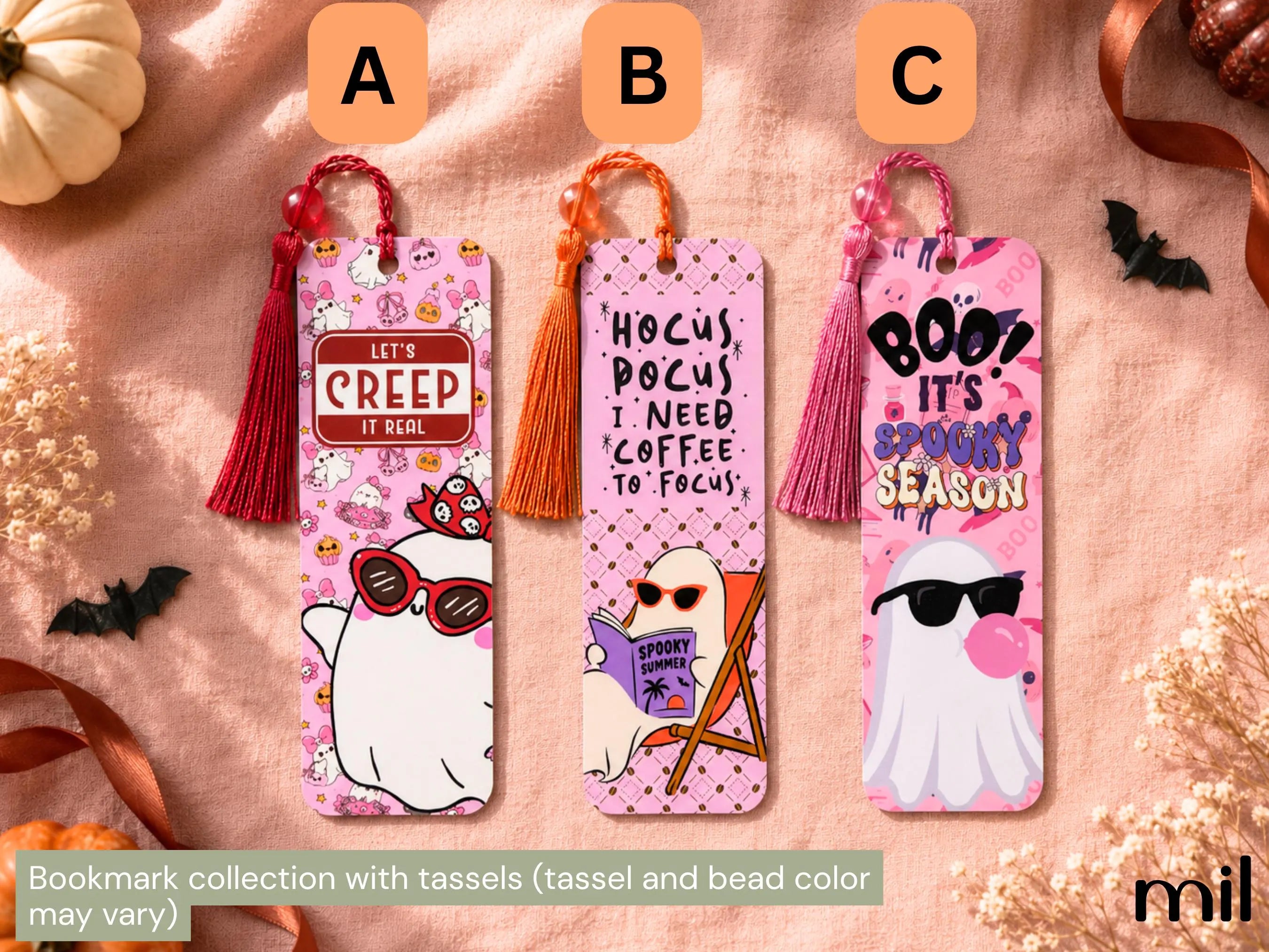 Three Halloween-themed bookmark sets with tassels are displayed on a textured pink fabric background. The bookmarks feature cute cartoon ghost illustrations with funny quotes: 'Let's CREEP IT REAL' on a red tassel, 'HOCUS POCUS I NEED COFFEE TO FOCUS' on an orange tassel, and 'BOO! IT'S SPOOKY SEASON' on a pink tassel.