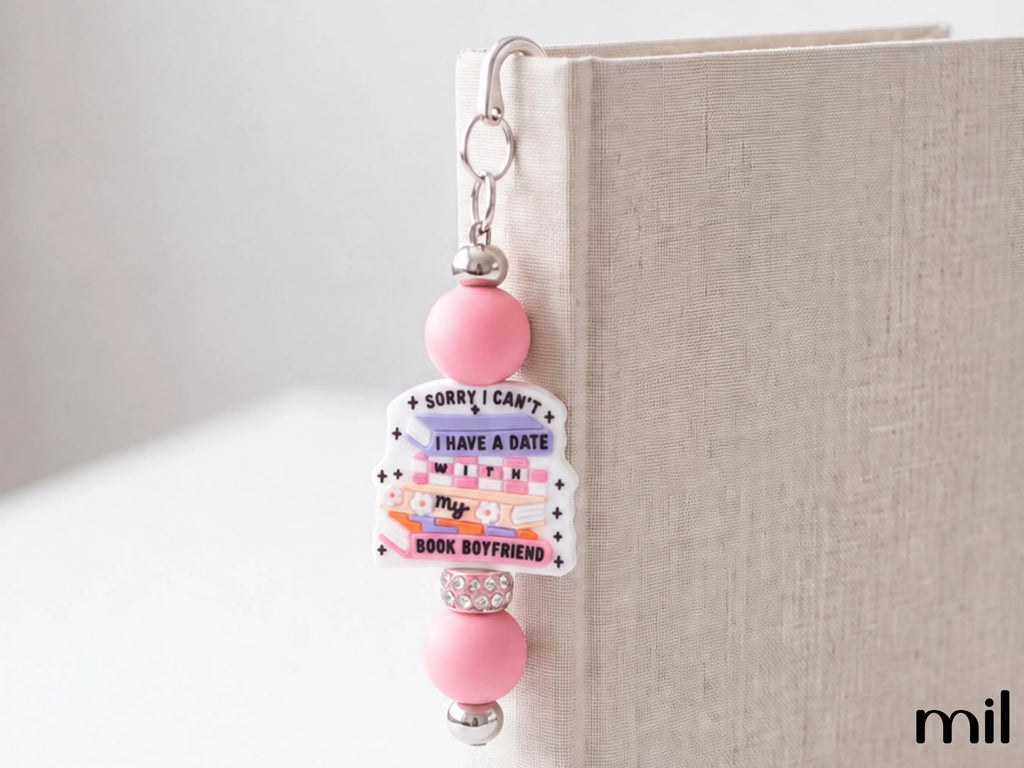A pink and white beaded book bookmark with the text 'SORRY I CAN'T I HAVE A DATE WITH MY BOOK BOYFRIEND' hanging from a silver metal clip on the spine of a beige book.