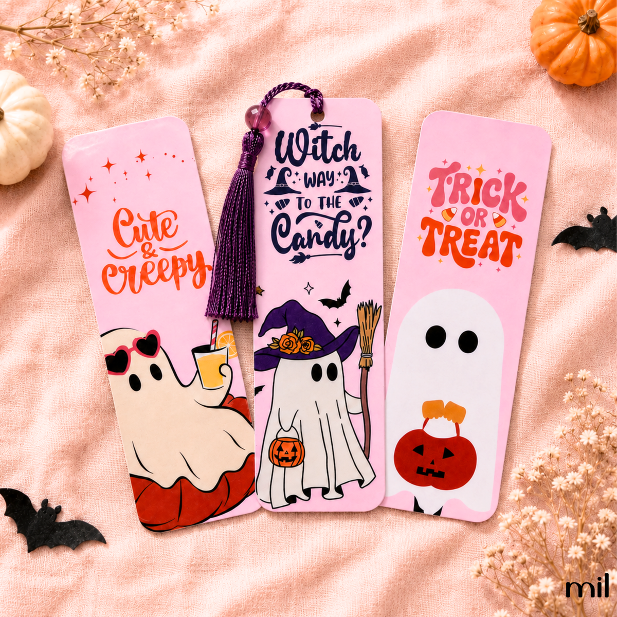 Three pink Halloween-themed metal bookmarks are arranged on a peach fabric background with decorative pumpkins, dried flowers, and black bat cutouts. The bookmarks feature whimsical illustrations and text: 'Cute & Creepy' with a ghost in sunglasses, 'Witch Way to the Candy?' with a witch in a hat, and 'Trick or Treat' with a ghost holding a pumpkin.