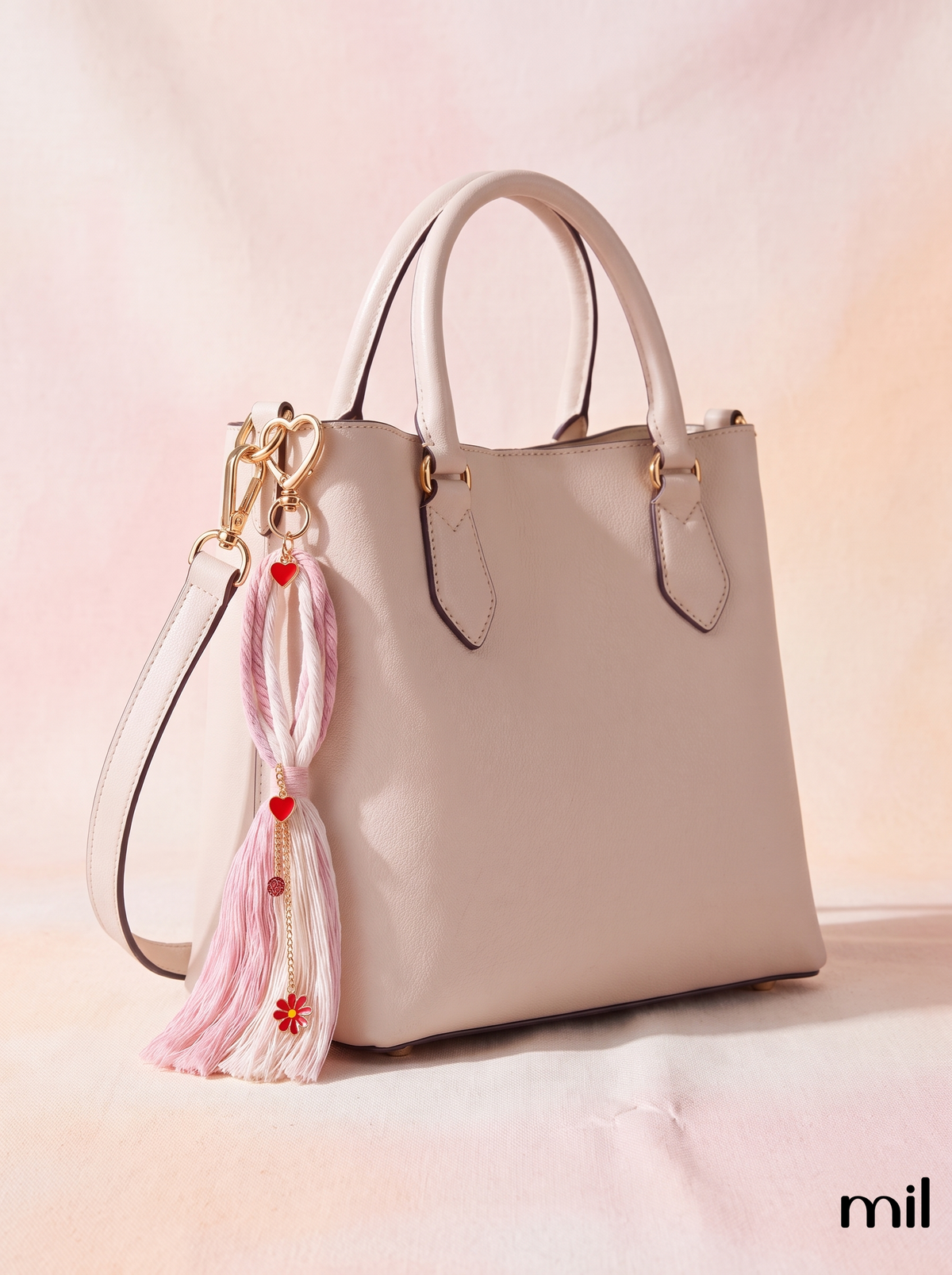 A light beige leather tote bag with double top handles, gold-tone hardware, and a decorative pink tassel keychain with red hearts and flowers hanging from the side, set against a soft pink background.