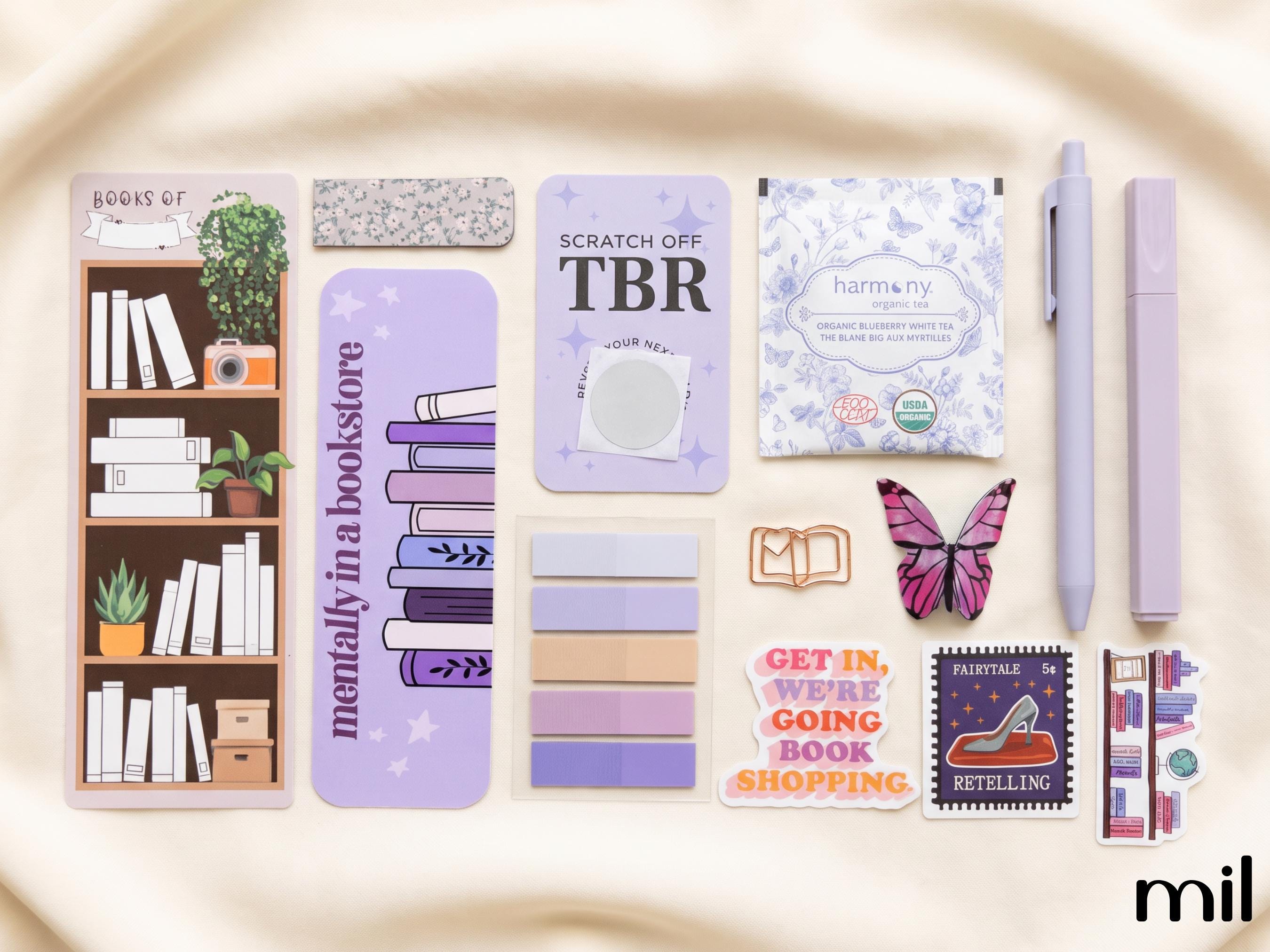A flat-lay arrangement of pastel purple and lavender stationery items on a cream fabric background, including a bookshelf planner page, sticky notes, a scratch-off note, a butterfly sticker, a pen, and other book-themed accessories.