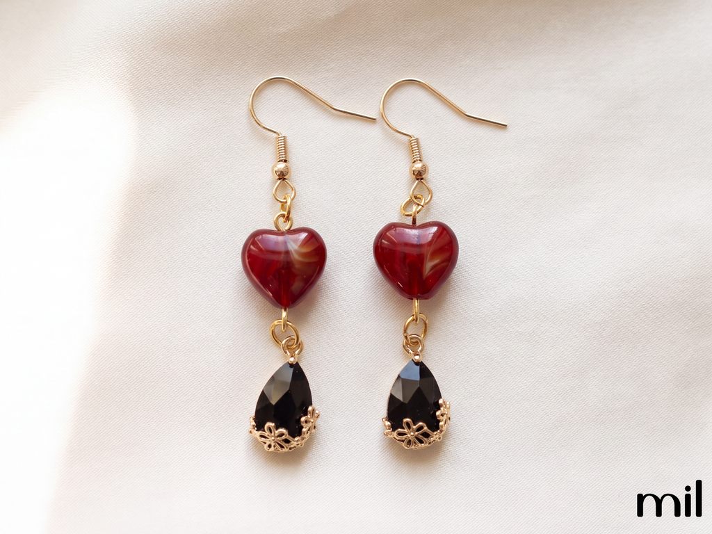 A pair of gold-tone dangle earrings featuring a polished dark red heart-shaped bead and a faceted black teardrop gemstone, displayed on a soft white fabric background.