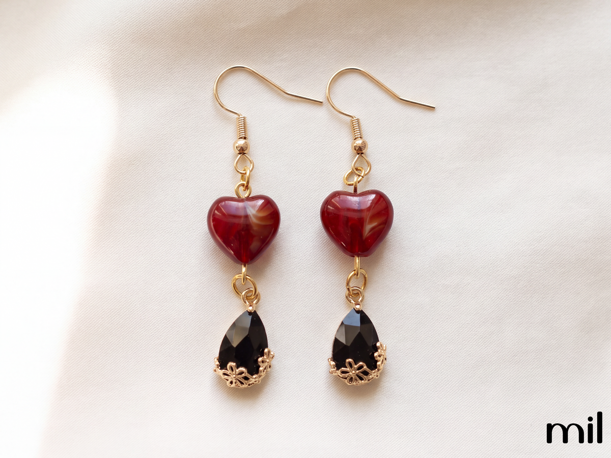 A pair of gold-tone dangle earrings featuring a polished dark red heart-shaped bead and a faceted black teardrop gemstone, displayed on a soft white fabric background.