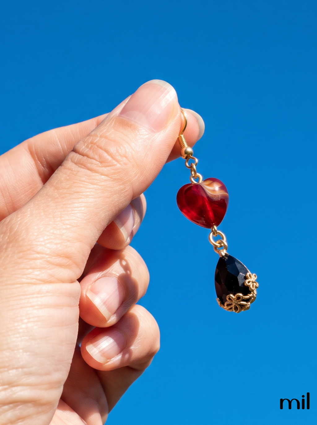 A hand holding a gold-tone dangle earring with a red heart-shaped crystal and a dark teardrop-shaped crystal against a bright blue sky.