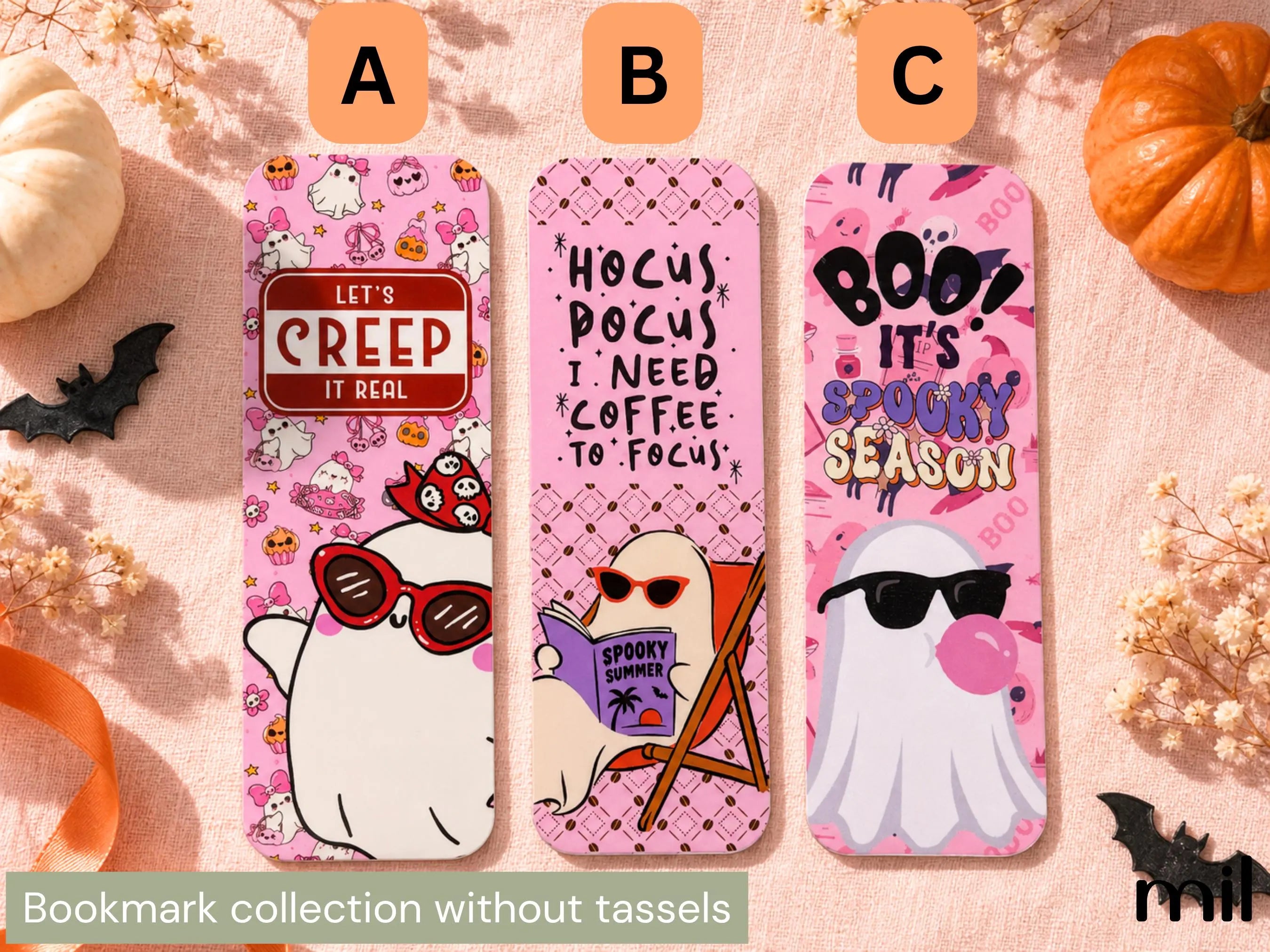 Three pink Halloween-themed bookmarks labeled A, B, and C are arranged on a peach fabric background with pumpkins, dried flowers, and black bat cutouts. The bookmarks feature cartoon ghosts, bold text, and seasonal graphics.