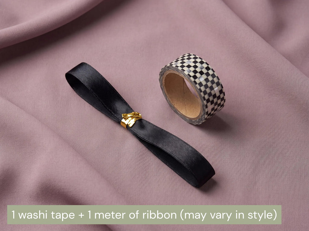 A black satin ribbon with a small gold decorative clasp and a roll of black-and-white checkered washi tape, arranged on draped dusty pink fabric. A text banner at the bottom reads: '1 washi tape + 1 meter of ribbon (may vary in style)'.