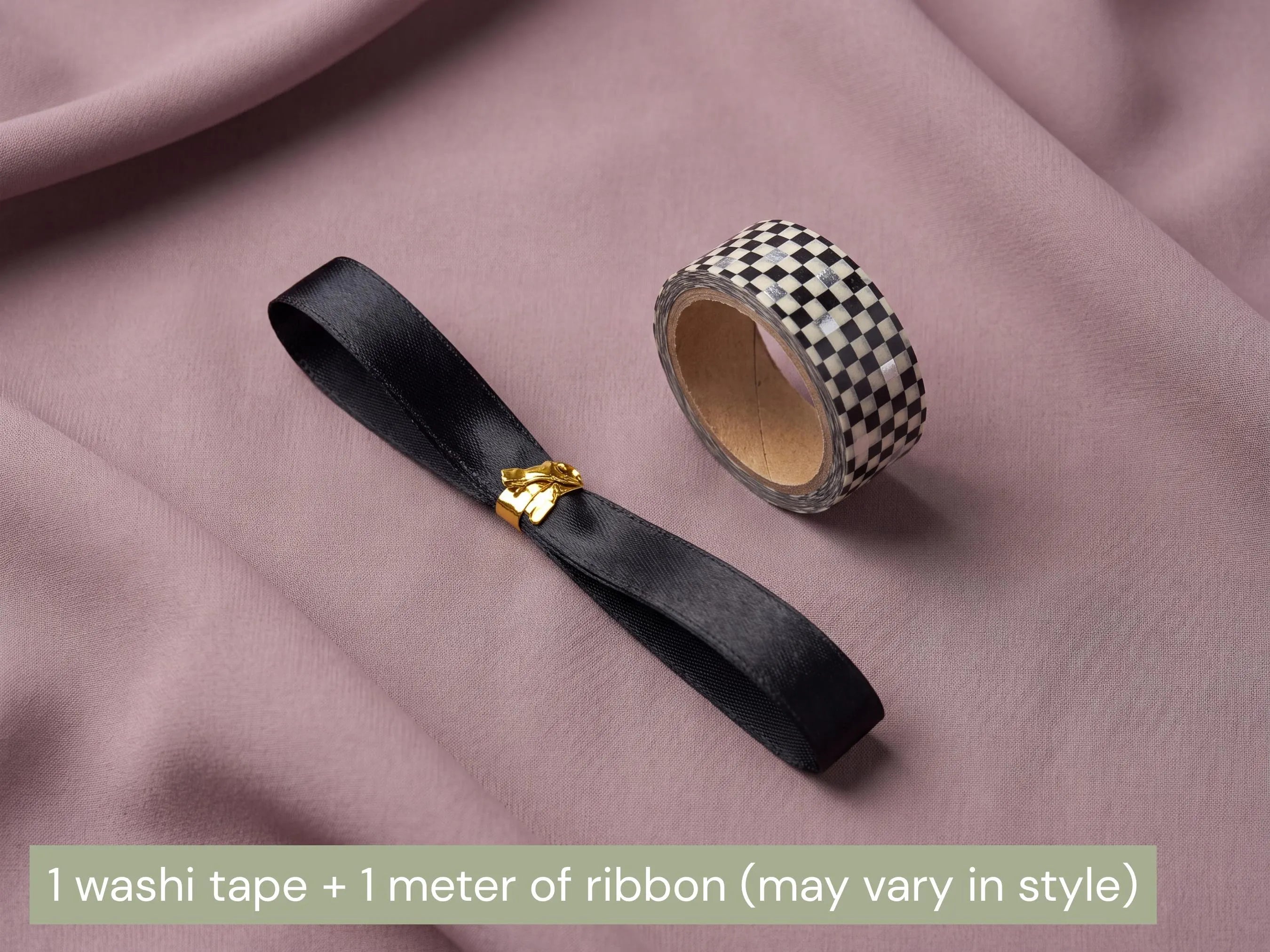 A black satin ribbon with a small gold decorative clasp and a roll of black-and-white checkered washi tape, arranged on draped dusty pink fabric. A text banner at the bottom reads: '1 washi tape + 1 meter of ribbon (may vary in style)'.