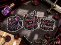 Three decorative stickers with gothic fantasy text are arranged on a dark tabletop with themed props including a small purple bottle, sword, velvet cloth, wax seal, chess piece, and rose petals. The stickers read: 'Here for the Magic and the Tension,' 'Give Me Swords and Yearning,' and 'Enemies to Lovers Enthusiast.
