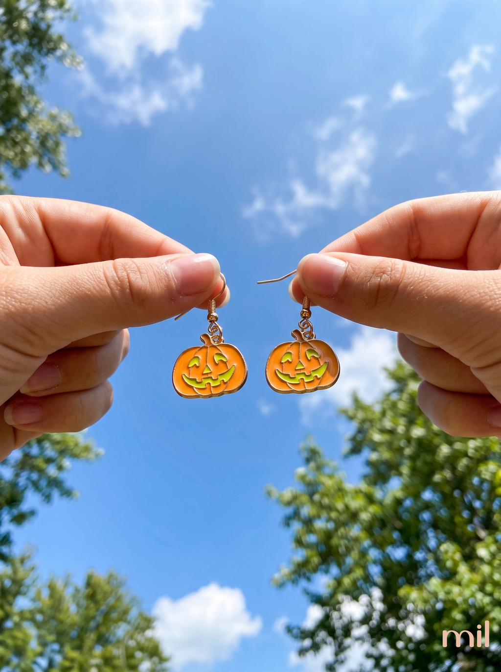 A person holds a pair of dangling earrings between two hands against a bright blue sky with clouds and tree foliage. The earrings are pumpkin-shaped with orange and yellow enamel detailing and small gold-toned hooks.