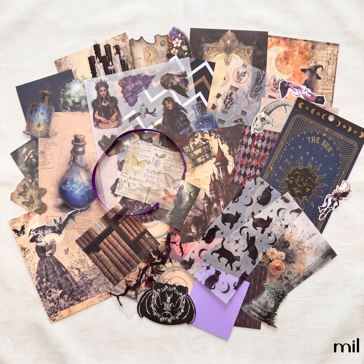 A flat-lay collage of vintage-style scrapbook papers, stickers, and decorative elements in muted sepia, black, cream, and purple tones, arranged on a light fabric background. A purple circular sticker or label is visible near the center, and the wordmark 'mil' appears in the bottom right corner.