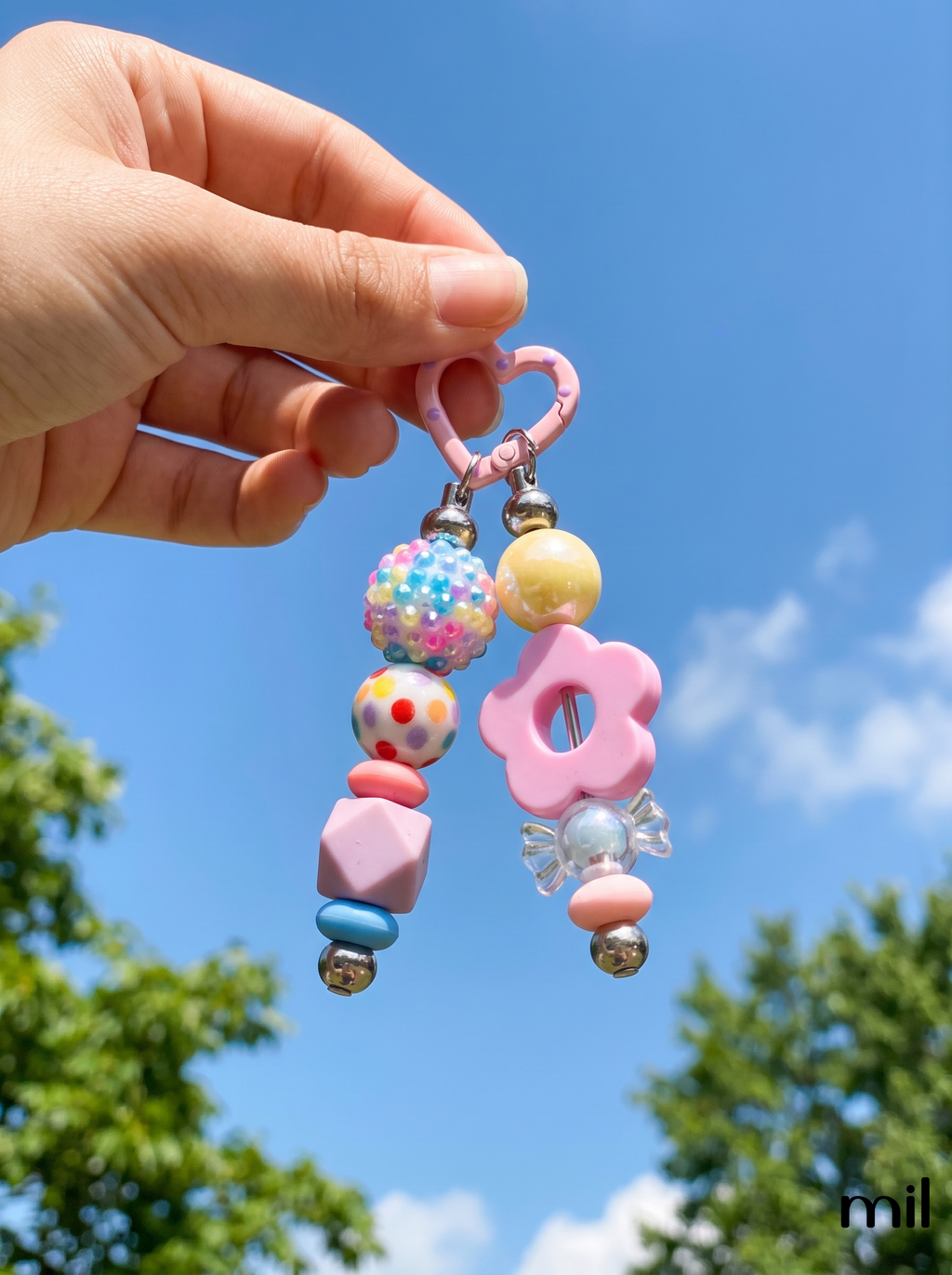 A hand holds a pastel pink beaded keychain outdoors against a bright blue sky with green trees. The keychain features a pink heart-shaped clasp, a pink flower charm, and assorted pastel beads including pink, yellow, blue, and white.